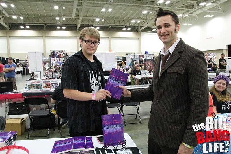 Robert Midgett handing a copy of Vagabonds to a man in a pinstripe suit at a convention table